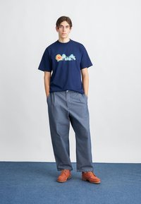 Kék alapú tengerészkék póló színes "Carhartt" logóval, laza világoskék farmernadrággal és narancssárga, texturált talpú cipőkkel egy kék padlón.