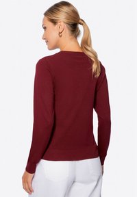 Maglione bordeaux a maniche lunghe con scollatura rotonda e polsini a costine. Texture a maglia con una silhouette aderente. Pantaloni bianchi visibili sotto la vita.
