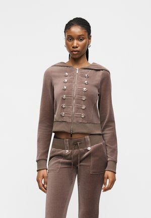 Nő fonott hajjal, barna cipzáras kapucnis pulóvert visel díszgombokkal és hozzáillő, zsinórral állítható nadrágot egyszínű háttér előtt.