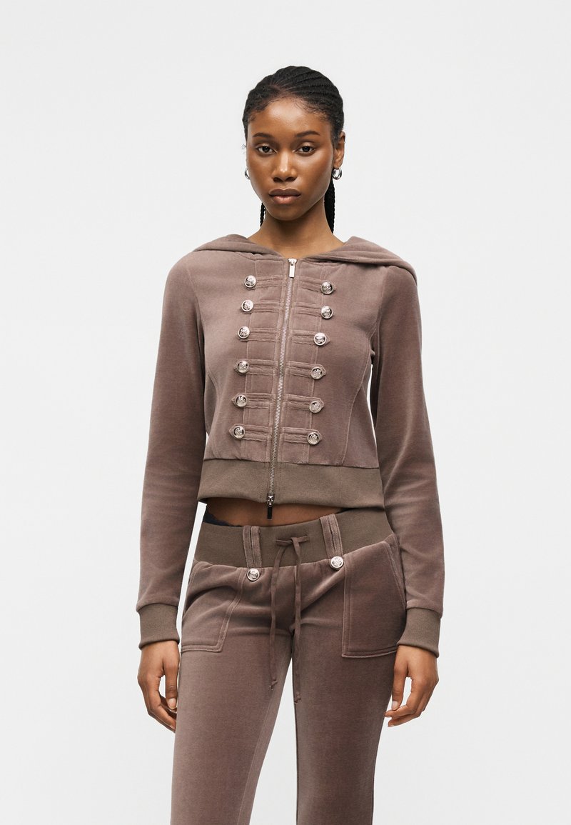 Kvinde med flettet hår iført en brun hættetrøje med lynlås og dekorative knapper samt matchende snørede bukser mod en ensfarvet baggrund.