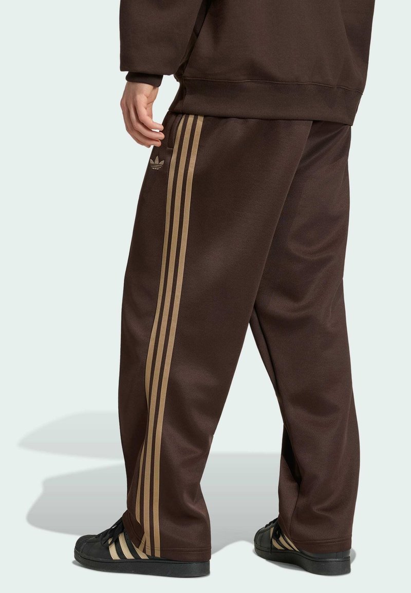 Person trägt braune Adidas-Trainingshose mit beigefarbenen Streifen und schwarzen Sneakers, gezeigt von der Rückenmitte bis zu den Füßen, steht auf hellem Hintergrund.