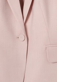 Blazer rose pâle avec un seul bouton recouvert, un revers cranté et une poche à rabat du côté droit.