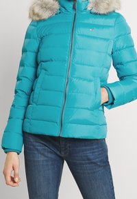 Turquoise donsjack met horizontale stiksels, ritssluiting en afneembare nepbontkraag. Voorzien van zijzakken en geribbelde manchetten.