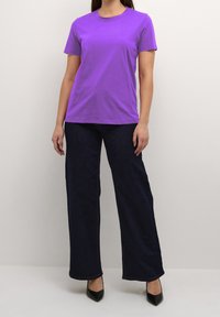 Kvinna iklädd en enkel lila t-shirt med korta ärmar, vida mörkblå jeans och svarta pumps med spetsiga tår, stående mot en ljus bakgrund.