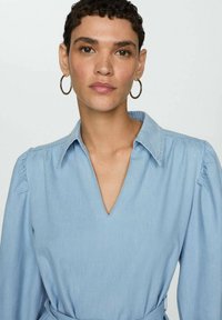 Robe en denim bleu clair avec un décolleté en V, des manches bouffantes et une taille à nouer. Texture lisse avec un design décontracté et sans motifs visibles.