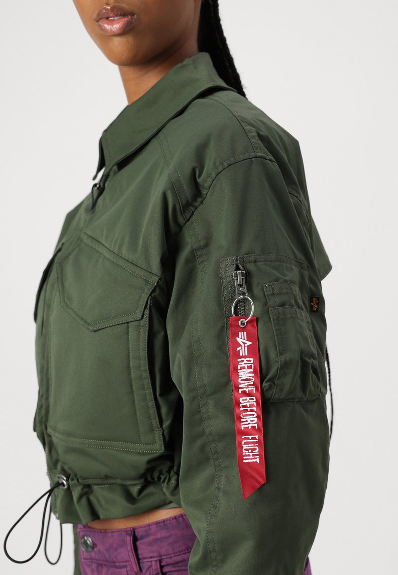 Chaqueta corta verde oliva con cremallera frontal, dos bolsillos en el pecho, cordón elástico en la cintura y una etiqueta roja "Retirar antes del vuelo".