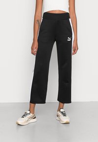 Pantalones de chándal negros con rayas laterales blancas y un logo de Puma. Cintura alta elástica, corte holgado y textura de tela suave. Combinados con zapatillas ligeras.