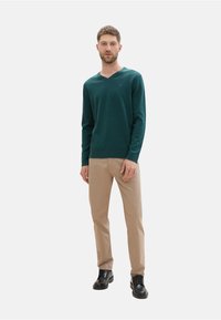 Pull vert en V, pantalons beiges, chaussures noires. Le pull a des manches longues, des poignets et un ourlet côtelés ; les pantalons sont droits avec une texture lisse.