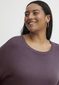 Lila pulóver széles nyakkivágással, puha, textúrált anyagból készült. A modellen arany fülbevalók és egyenes, sötét haj látható.
