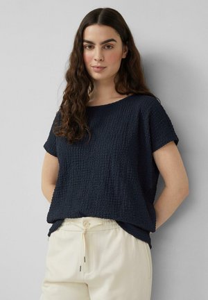Vrouw met lang, krullend bruin haar, gekleed in een navy textuur top met korte mouwen en crème broek met trekkoord, tegen een effen grijze achtergrond.