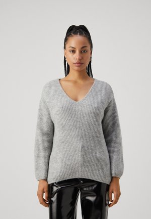 Pull en maille grise avec un col en V, un design texturé et des manches longues, associé à un pantalon noir brillant.