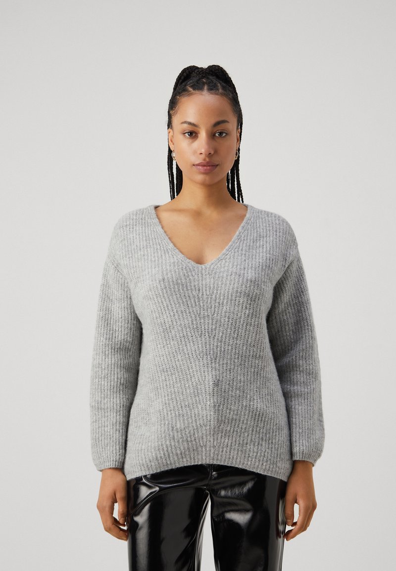 Grauer gestrickter Pullover mit V-Ausschnitt, strukturiertem Design und langen Ärmeln, kombiniert mit glänzenden schwarzen Hosen.