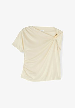 Kremowa bluzka z krótkim rękawem z asymetrycznym dekoltem, marszczona i zabezpieczona złotą prostokątną klamrą na prawym ramieniu.