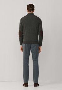 Donkergroene gebreide trui met een hoge kraag en bruine elleboogpatches, gecombineerd met lichtgrijze broeken en donkeren bruine schoenen.