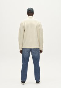 Beige vlněná bunda s límcem, bočními rozparky a knoflíkovými manžetami, oblečená přes modré džíny. Model nosí zelenou čepici.