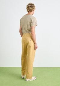 Chaki medvilniniai platūs kelnės su dviem užpakalinėmis kišenėmis, subtiliais siuvimo akcentais ir Carhartt logotipo juostele. Dėvimos su smėlio spalvos marškinėliais ir baltais sportiniais bateliais.