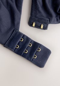 Navy mėlynos spalvos audinio drabužis su lygiu paviršiumi, turintis auksinės spalvos metalines detales ir elastines petnešėles. Komplekte yra keletas kilpų užsegimų reguliavimui.