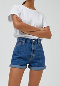 Shorts de mezclilla en azul medio con puños enrollados, cintura alta y cinco bolsillos. Combinados con una camiseta blanca de cuello redondo sin estampado.
