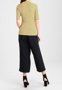 Top de manga corta a rayas amarillas y blancas combinado con pantalones de pierna ancha en color azul marino y zapatos de tacón bloque negro. Vista trasera, textura de tela suave.