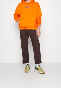 Sudadera naranja con capucha, pantalones marrones con puños y zapatillas multicolores con acentos verdes y amarillos. Atuendo urbano casual.