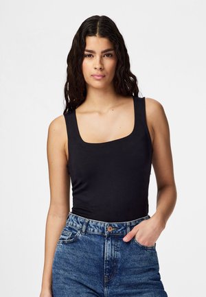 Donna con lunghi capelli scuri e ricci, indossa un top nero senza maniche e jeans blu a vita alta, con una mano in tasca, su sfondo bianco.