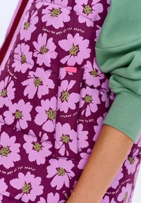 Kurtka pikowana w kolorze fioletowym z dużymi różowymi kwiatami i zielonymi środkami, z boczną kieszenią i napisem, noszona z zieloną koszulą z długim rękawem.