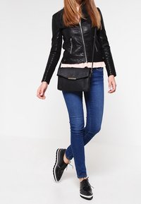 Donna che indossa una giacca di pelle nera, jeans skinny blu, scarpe con piattaforma nere con lacci e una borsa a tracolla nera, in piedi contro uno sfondo bianco.