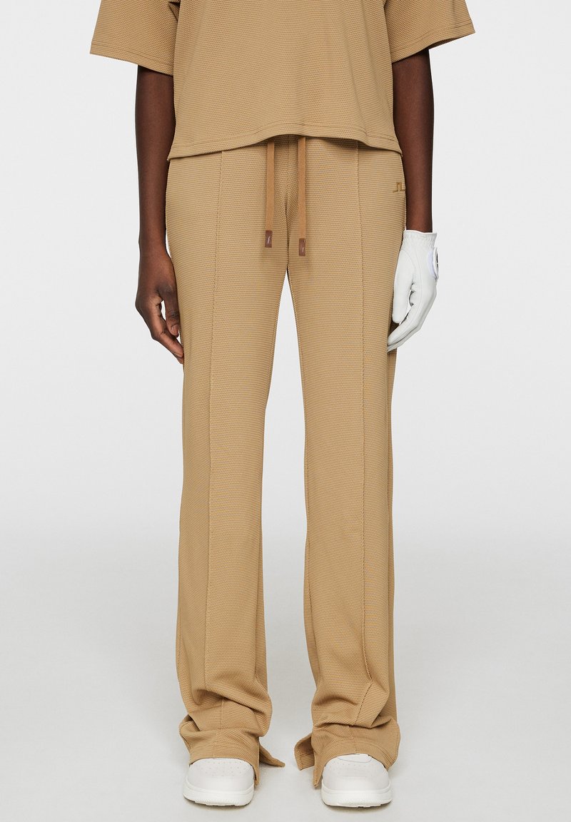 Beige Stoffhosen mit lockerer Passform, ausgestattet mit einem Kordelzugbund, betonten Nähten und kombiniert mit weißen Turnschuhen.