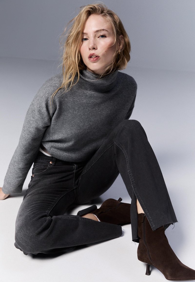 Szary sweter w golfie, ciemnoszare dżinsy z surowymi brzegami oraz brązowe zamszowe botki na kostkę z małym obcasem. Miękkie tekstury, dopasowany krój.