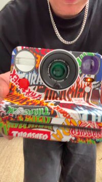 Polaroid-kamera med et fargerikt, grafisk design med ulike mønstre og logoer, inkludert flammer og ordet "THRASHED," sirkulær linse, og blits.
