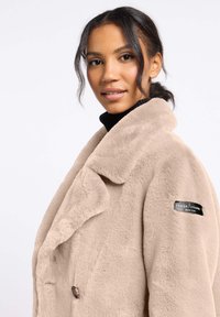 Beige nep-bontjas met brede kraag, oversized pasvorm, dubbele knoopsluiting en logo patch op de mouw. Zachte textuur en luxueuze afwerking.