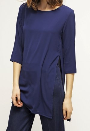 Tunique bleu marine à manches longues en tissu lisse, avec un col rond et des fentes sur les côtés pour un ajustement décontracté, associée à un pantalon sombre.
