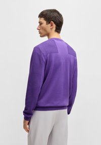 Lila tröja med ribbad textur på ärmarna och bakpanelen. Rundhalsad design med avslappnad passform, kombinerad med ljusa byxor.