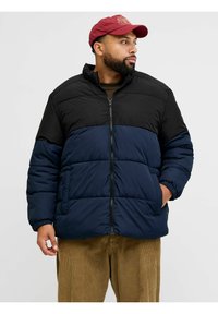 Gepolsterte Jacke mit schwarzem Oberteil und marineblauem Unterteil. Reißverschluss, hoher Kragen und elastische Bündchen. Getragen über beigen Cordsamt-Hosen.