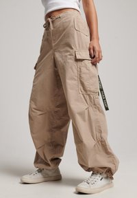 Beige cargo byxor med en avslappnad passform, som har flera fickor, dragsko i midjan och cuffs nederst; tillverkade av ett lätt material.