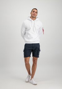 Biely sveter s kapucňou a prednou vreckom, skombinovaný s námorníckymi cargo šortkami s bočnými vreckami. Biely tenisový obuv dokončuje outfit.