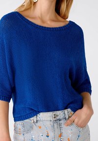 Niebieski sweter dzianinowy z szerokim dekoltem i krótkimi rękawami, ozdobiony teksturowym materiałem, w połączeniu z jasnoniebieskimi dżinsami o wzorze z plam farby.