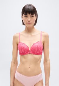 Γυναίκα με κοντά σκούρα μαλλιά που φοράει ροζ δαντελωτό σουτιέν και ανοιχτό ροζ εσώρουχα με δαντέλα, μπροστά σε απλό ανοιχτό φόντο.