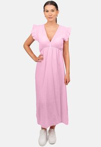 Roze, mouwloze maxi-jurk van getextureerde stof, met ruches op de schouders en een diepe V-hals. Gekleed met witte sneakers.
