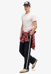 Camiseta blanca, camisa de cuadros roja atada a la cintura, joggers azul marino con franjas laterales y zapatillas cremosas con suelas de goma oscura.