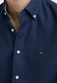 Primer plano de una persona que lleva una camisa de botones azul marino con botones blancos y un pequeño logotipo bordado en rojo, blanco y azul en el pecho.