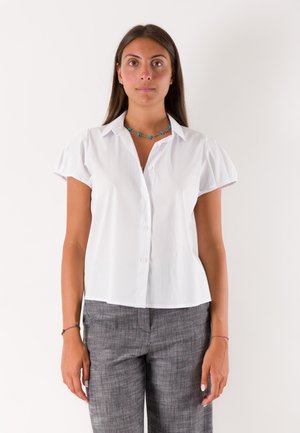 Giovane donna con lunghi capelli castani che indossa una camicia bianca a maniche corte con bottoni, pantaloni grigi e una collana turchese, in piedi davanti a uno sfondo bianco.