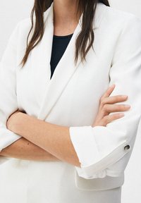 Blazer blanco con un diseño estructurado, que presenta mangas enrolladas con acentos de botón. Debajo, se puede ver una camiseta de punto negra.