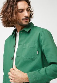 Groene, gekraagde jas van textuurstof met zwarte knopen, borstzak en label. Licht getailleerd ontwerp met lange mouwen.