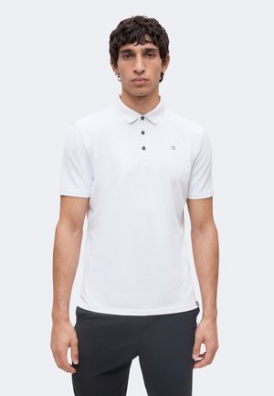 Giovane uomo con capelli scuri e ricci che indossa una polo bianca a maniche corte con tre bottoni e pantaloni neri su uno sfondo neutro.