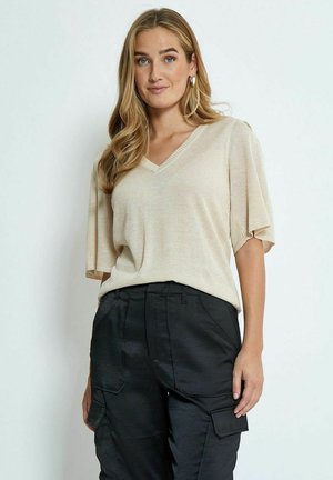 Beiger V-Ausschnitt-Pullover mit kurzen Ärmeln, aus einem weichen, texturierten Stoff gefertigt. Kombiniert mit schwarzen Cargo-Hosen mit Seitentaschen.
