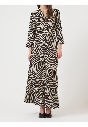 Frau trägt ein langes, geknöpftes Zebrakleid mit Dreiviertelärmeln und steht vor einem einfarbigen hellen Hintergrund.