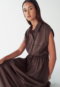 Hnedé ľanové šaty s krátkymi rukávmi, gombíkovou prednou časťou, dvomi náprsními vreckami a sťahovacím pásom, s voľným sukňou a jemnou textúrou.