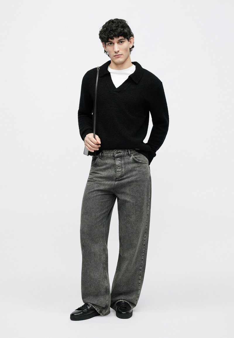 Young man with curly hair wearing black sweater, white undershirt, loose gray jeans, black sneakers, and carrying small black shoulder bag.