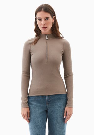 Mujer vestida con una camiseta de manga larga ajustada de canalé beige con cremallera delantera, combinada con jeans azules, de pie contra un fondo blanco.
